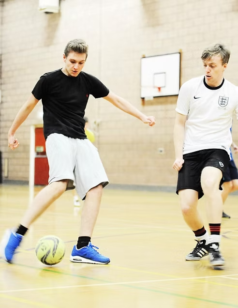 One young man kicking a football with a second young man attempting a tackle in a sports hall