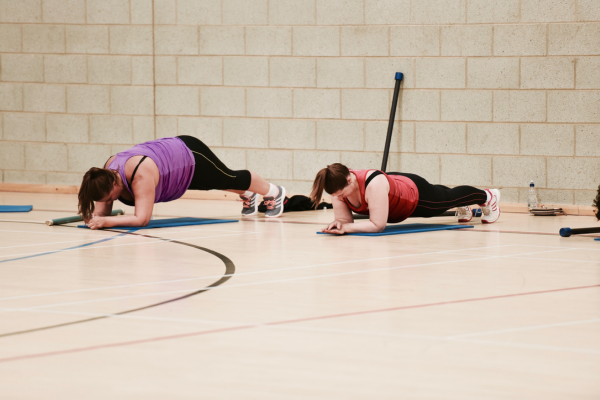 Two women in a plank position in a exercise class. One woman in wearing a purple vest top with black leggings and the other woman is wearing a red vest top with black leggings.