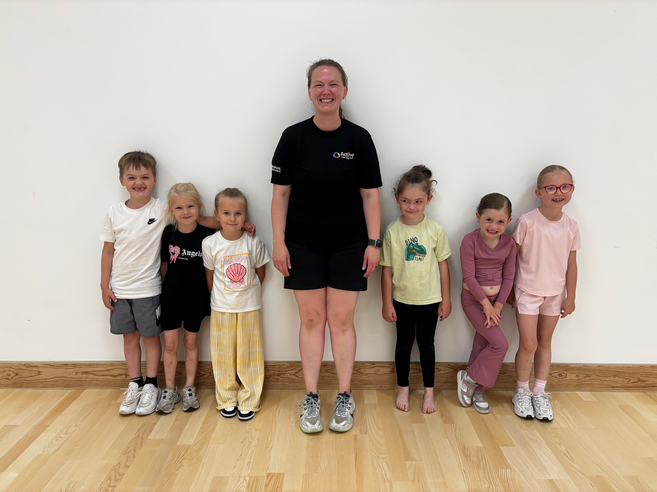 Active Hartlepool instructor wearing black shorts and a black tshirt with 3 children either side of her leaning against a white wall after their fitness class