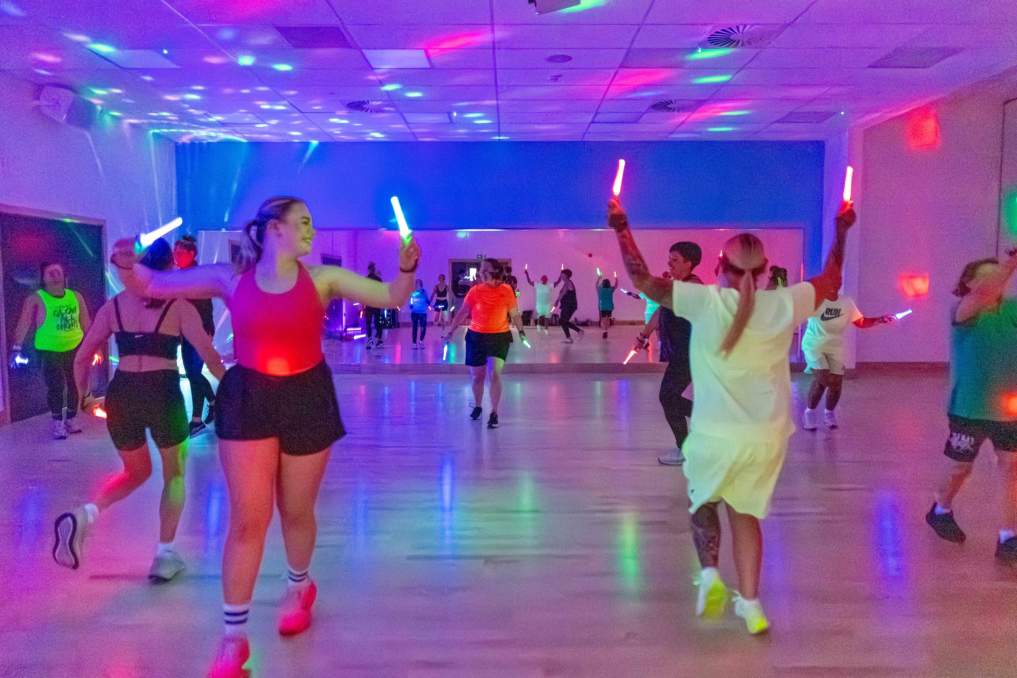 Photograph shows a Clubbercise class with people dancing with glow sticks in a room with strobe lights