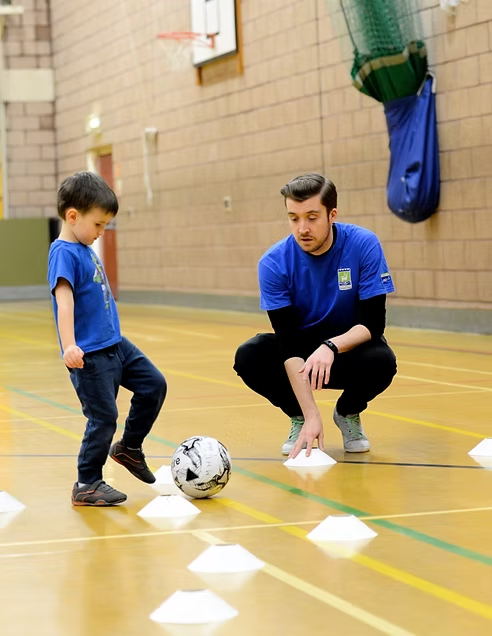 Male instructor and young boy dribbling football around plastic white cones in a sports hall