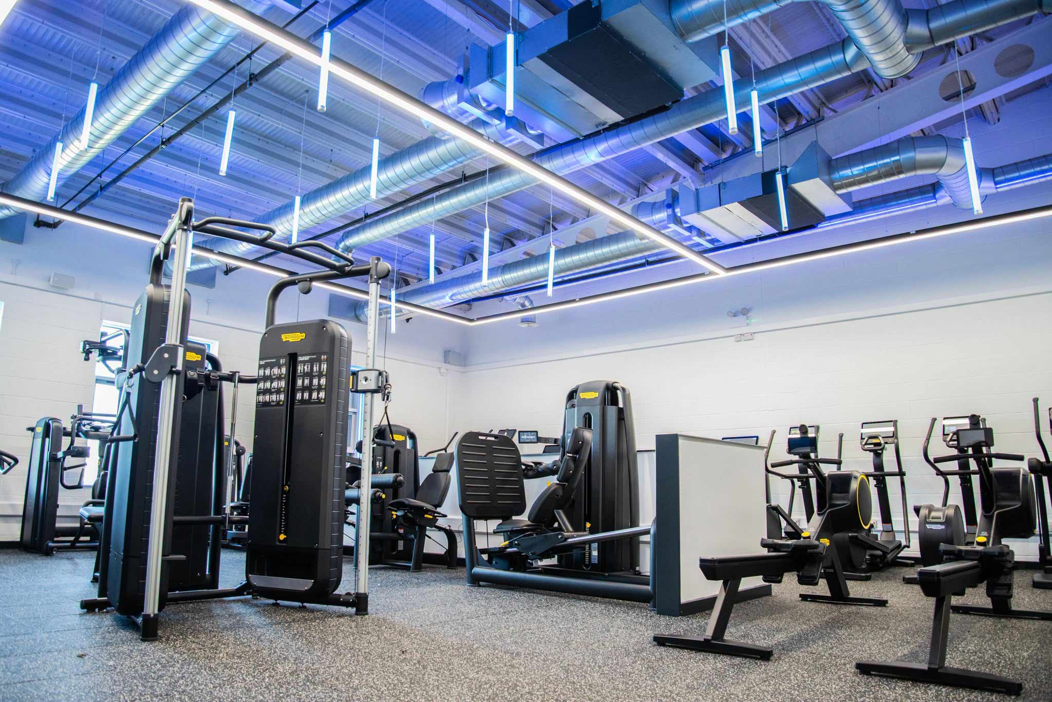 Photograph of the gym at Brierton Sports Centre that shows black Technogym cardio and weight equipment. The gym has a grey floor, white walls and blue LED ceiling lights