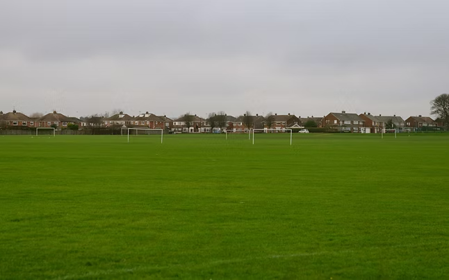 outdoor grassy field with multiple goal posts and visible houses in the distance